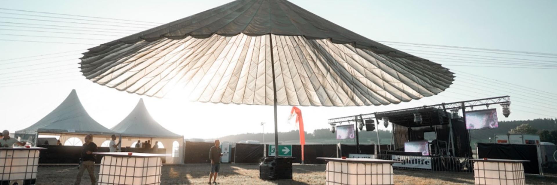 Das Bild zeigt eine Outdoor-Veranstaltung mit einem großen, schirmartigen Sonnenschutz aus Fallschirmstoff in der Mitte. Im Hintergrund sind zwei weiße Zelte mit spitzen Dächern und eine Bühne mit Bildschirmen und Beleuchtung zu sehen. Einige Menschen stehen im Bereich unter dem Fallschirm und bei Stehtischen. Der Himmel ist klar und sonnig.