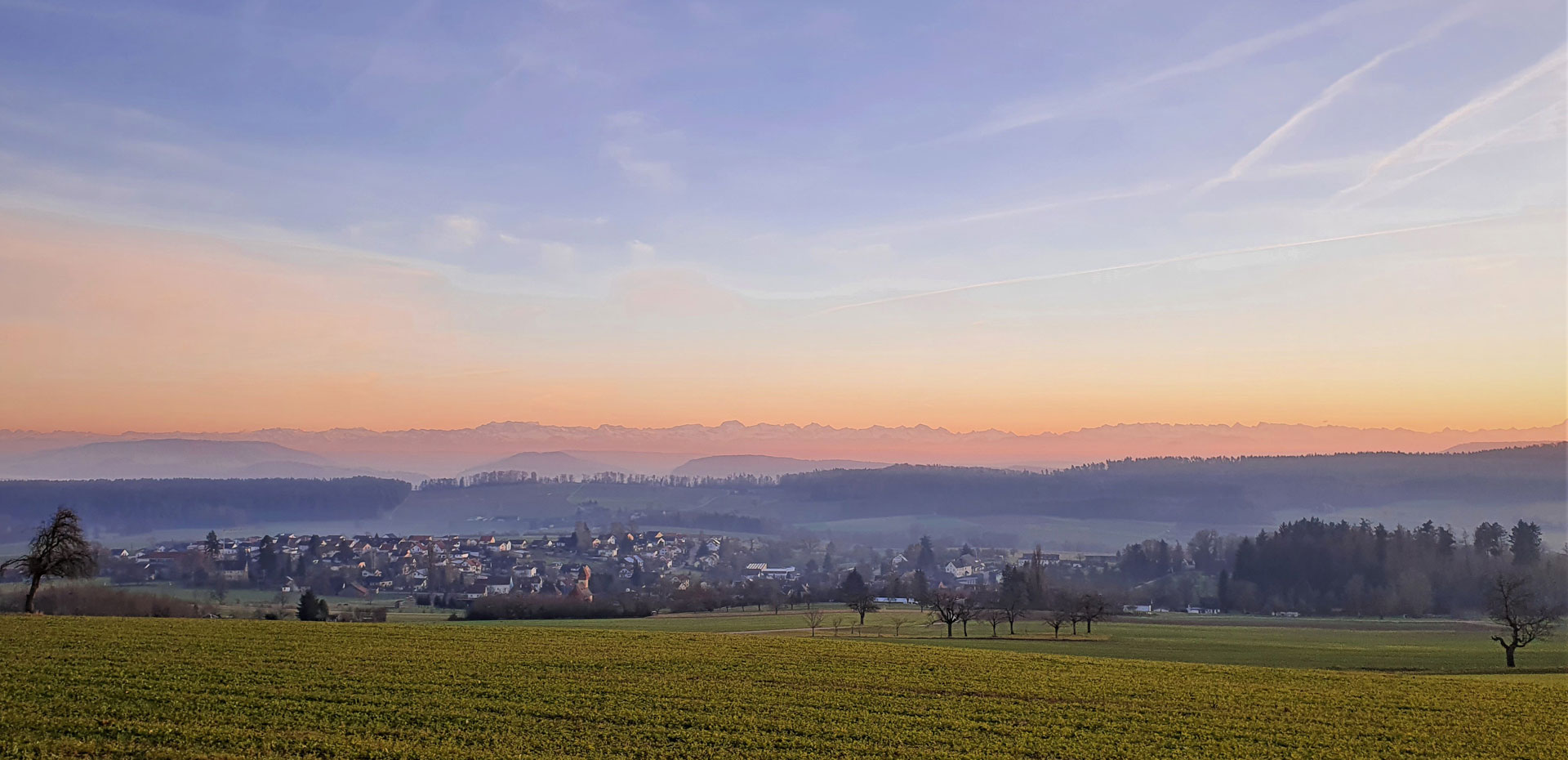 Blick auf Dettighofen und die Alpen bei Sonnenuntergang, die warmen Farben des Himmels reflektieren die ruhige Landschaft.